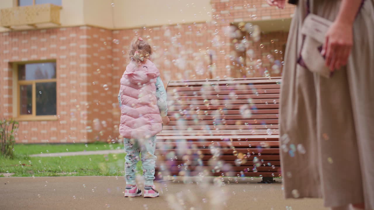 Girl in pink vest stands in courtyard watching rising soap bubbles, hands open with curiosity, adult nearby observes, bench and brick building behind, gentle outdoor play filled with discovery