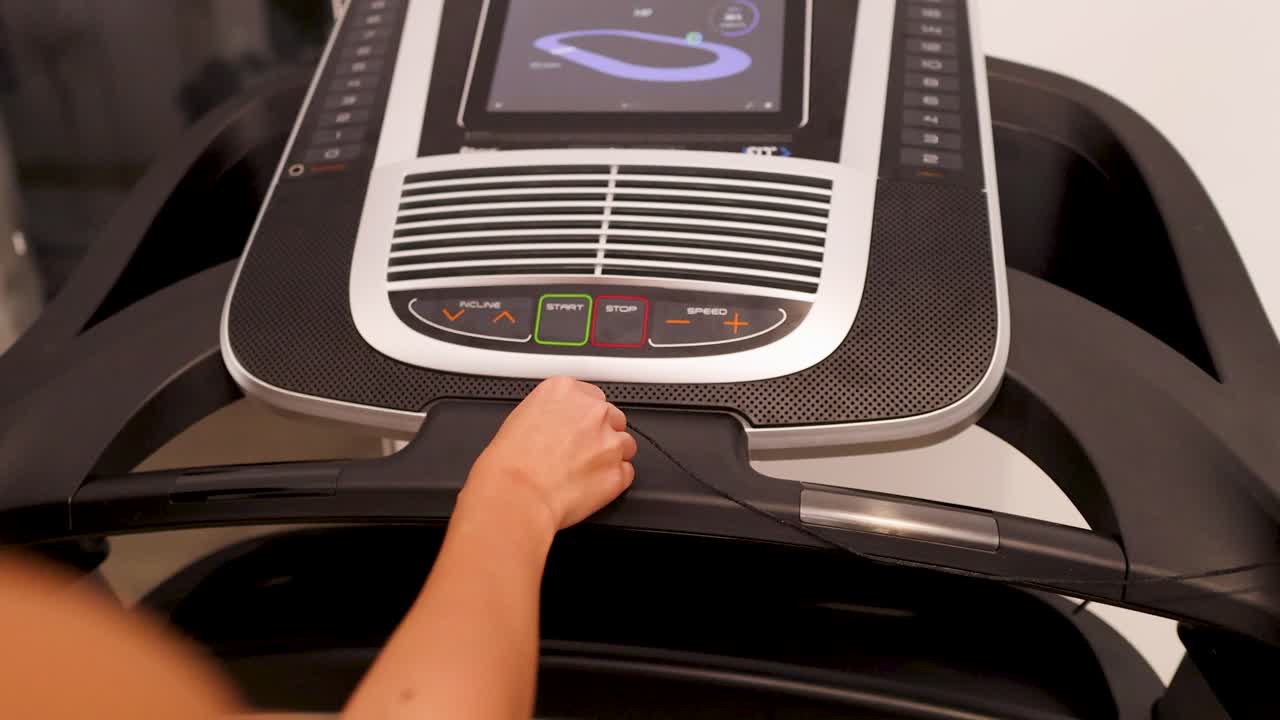 A woman interacts with a treadmill control panel in a home gym setting, adjusting settings under warm indoor lighting