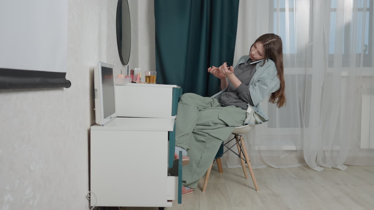 Donna caucasica che si rilassa sulla sedia della toeletta, luce naturale soffusa, tenda verde, cassettiera con boccette di smalto, estetista a domicilio seduta che si gode un tranquillo momento di cura di sé, sorriso delicato, atmosfera interna accogliente