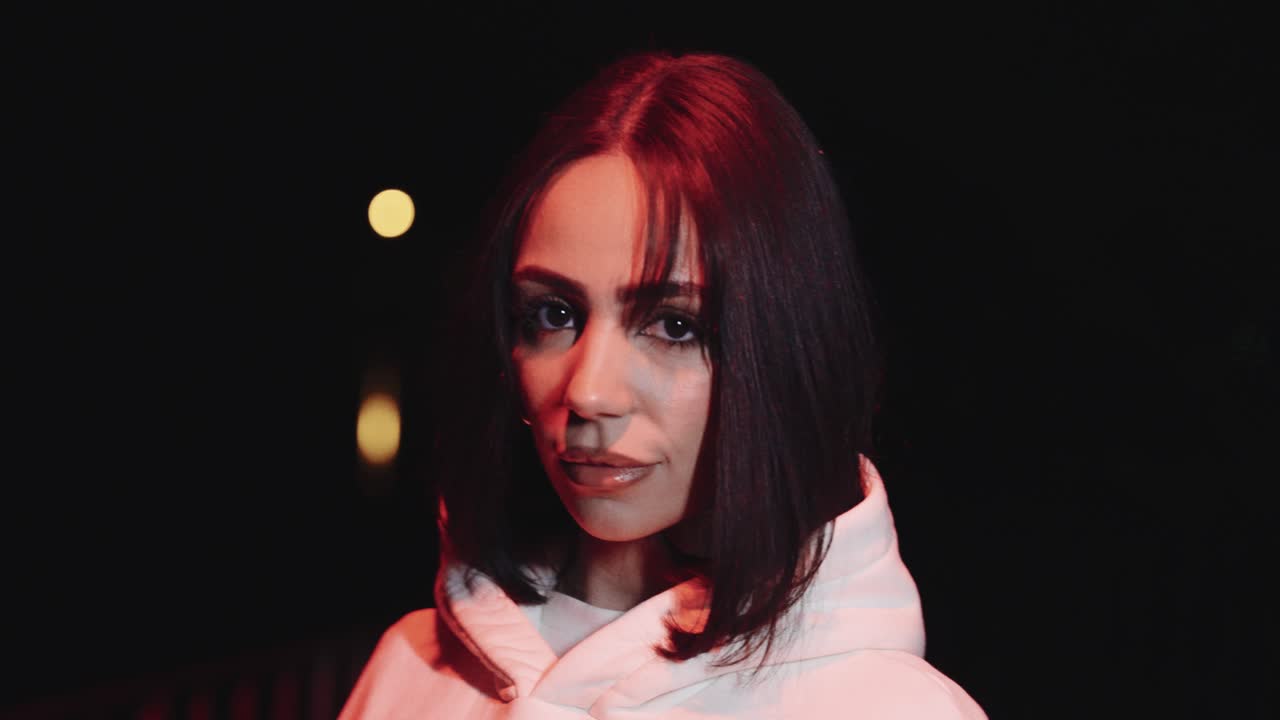 Young beautiful woman with white sweater. in the night looks into the camera, dark surroundings, poses for the camera and puts her hair back, close up