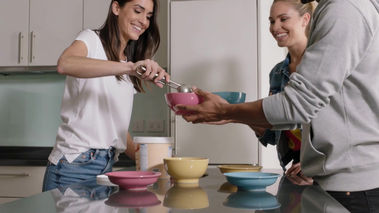 Mujer con camiseta blanca sirviendo de un recipiente grande a un tazón rosa, con amigos sosteniendo tazones en la isla de la cocina.
