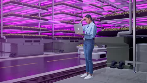 vista lateral completa del cuerpo de una trabajadora profesional asiática de pie con su computadora portátil en una granja vertical, instalación industrial de alta tecnología, escribiendo y decepcionada con asintiendo con la cabeza