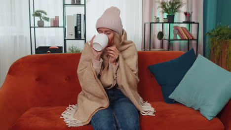 Sick-young-woman-wear-hat-wrapped-in-plaid-sit-alone-shivering-from-cold-on-sofa-drinking-hot-tea