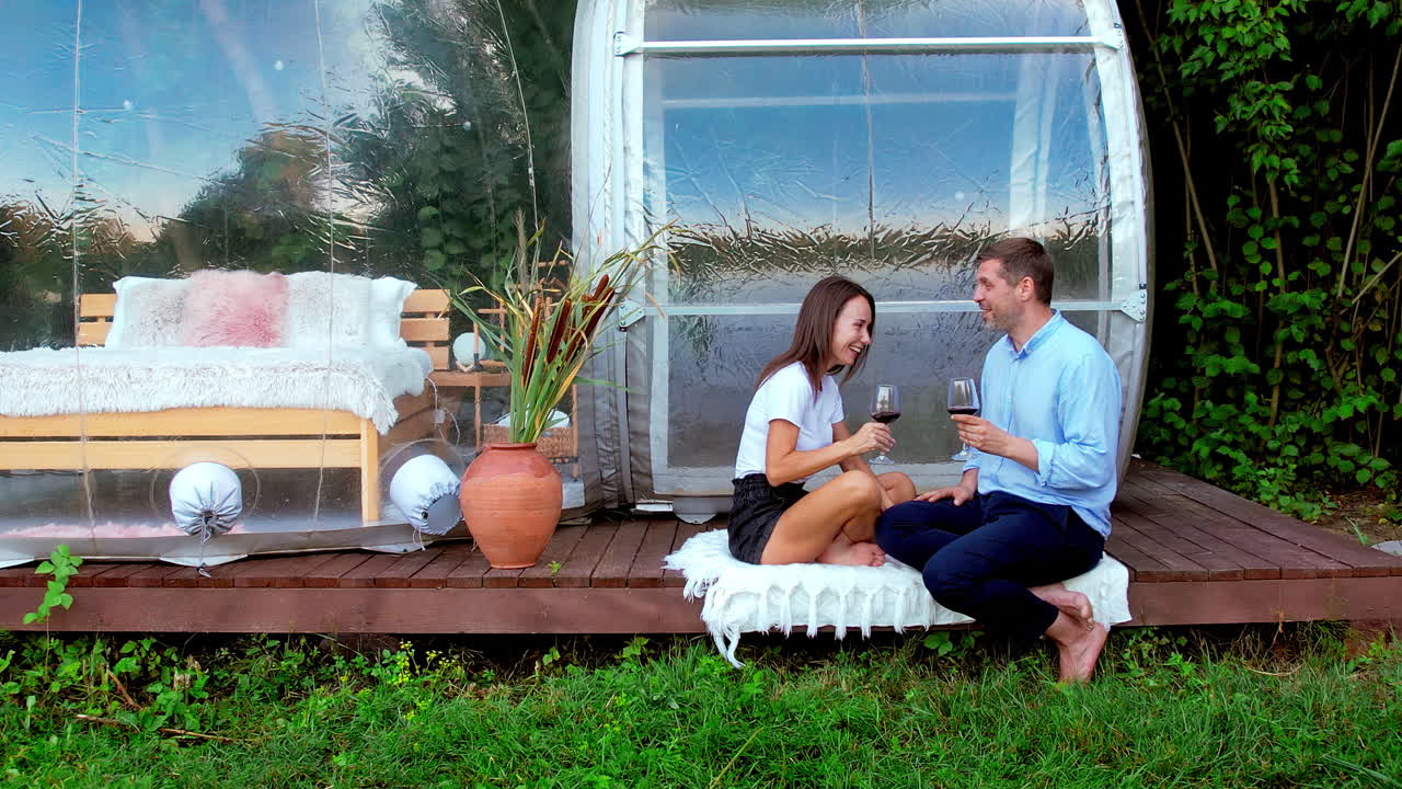 Happy couple drinking wine near transparent bubble tent at glamping. Nature around
