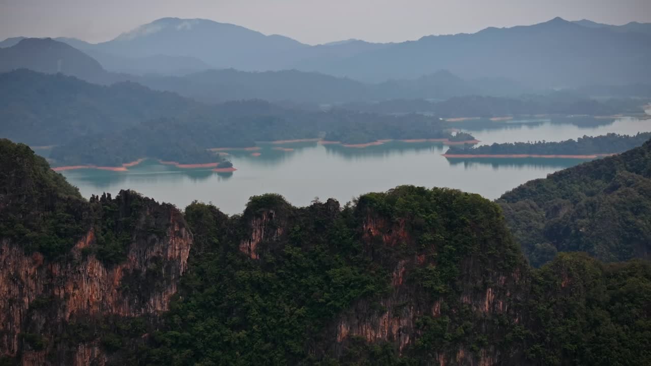Drone footage flying over limestone cliffs in Khao Sok National Park, Thailand, revealing rainforest, lake islands, and misty mountains in the distance