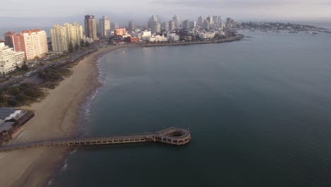 Luftaufnahme-Mit-Kreisförmiger-Anlegestelle-Und-Sandstrand-Mit-Skyline-Von-Punta-Del-Este-Im-Hintergrund-Während-Bewölkter-Tage