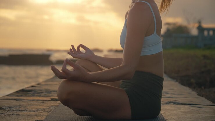 Mujer atlética y atractiva sentada en el suelo haciendo meditación de yoga con la luz del sol escénica de fondo, brisa en el cabello rubio, costa de Bali.