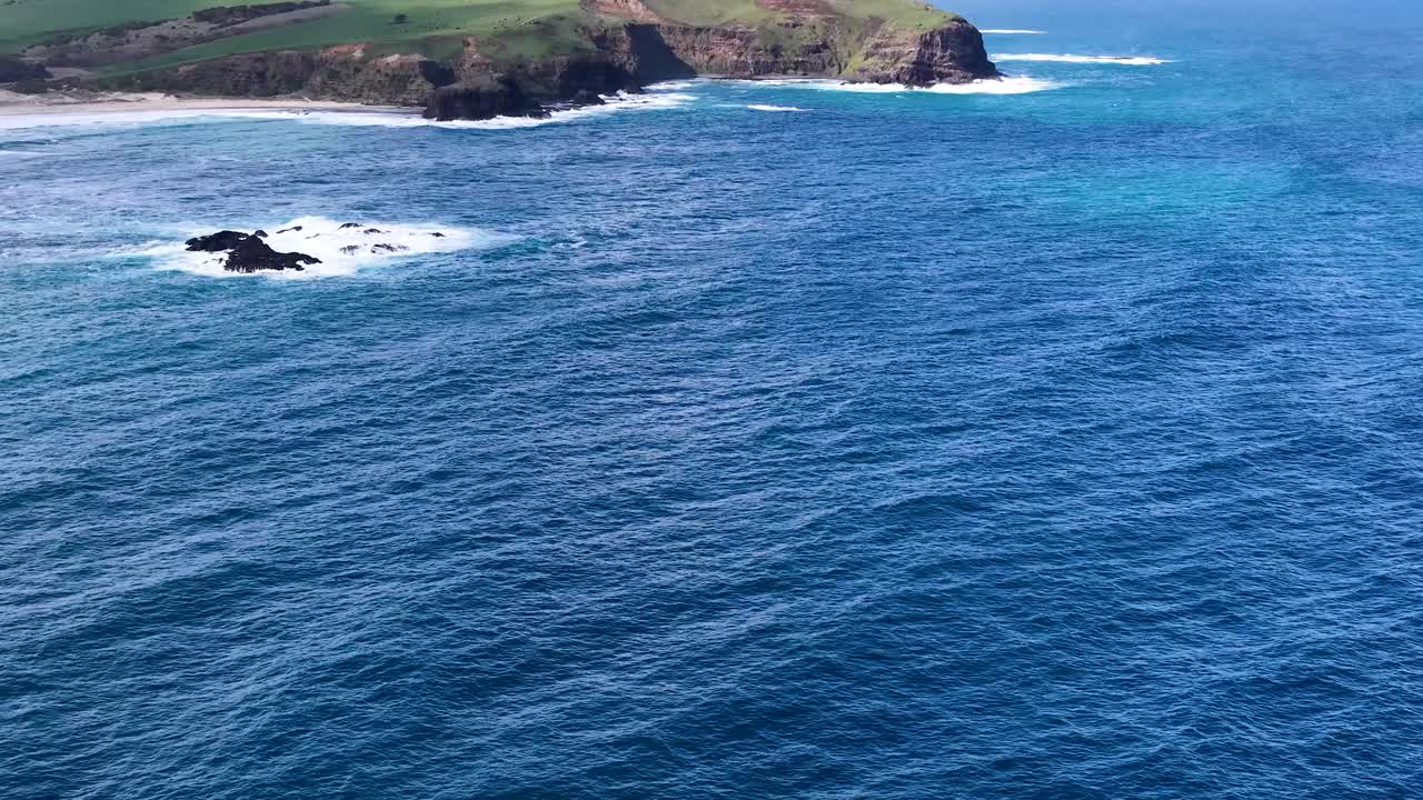 Drone camera glides over blue ocean, revealing rocky outcrop and rugged coastline in daylight
