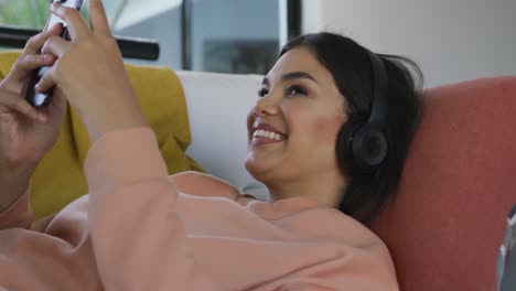 Video-Einer-Glücklichen-Gemischtrassigen-Frau,-Die-Mit-Kopfhörern-Auf-Dem-Sofa-Liegt-Und-Ihr-Smartphone-Benutzt