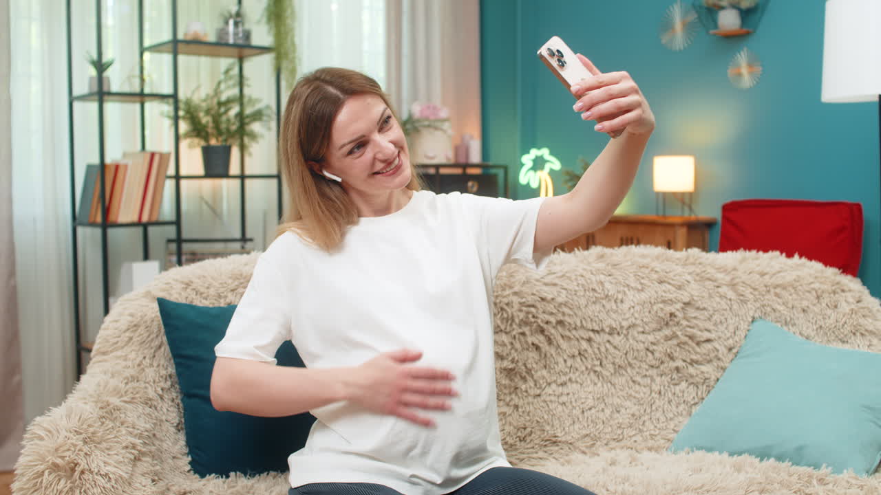 Pregnant woman on smartphone video call wearing earbuds waves hello shows belly happily with family