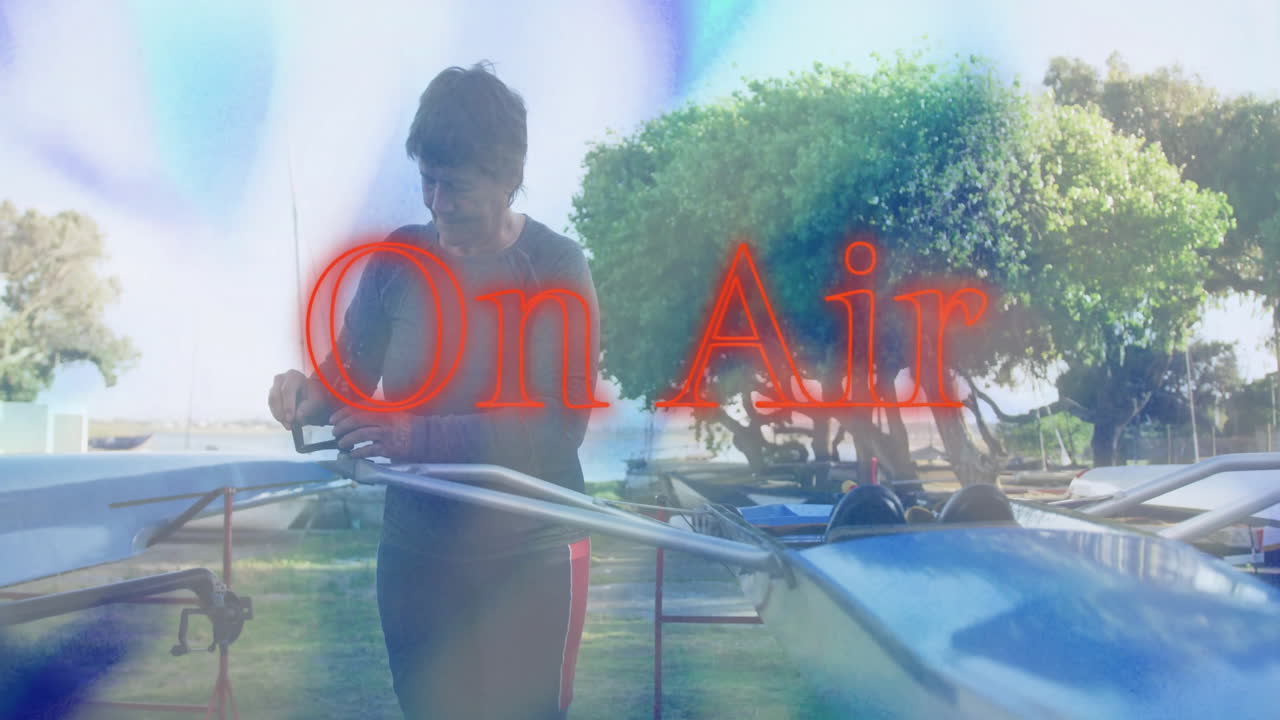 Senior woman adjusting rowing shell rigging in boatyard, showcasing broadcast tech On Air graphic