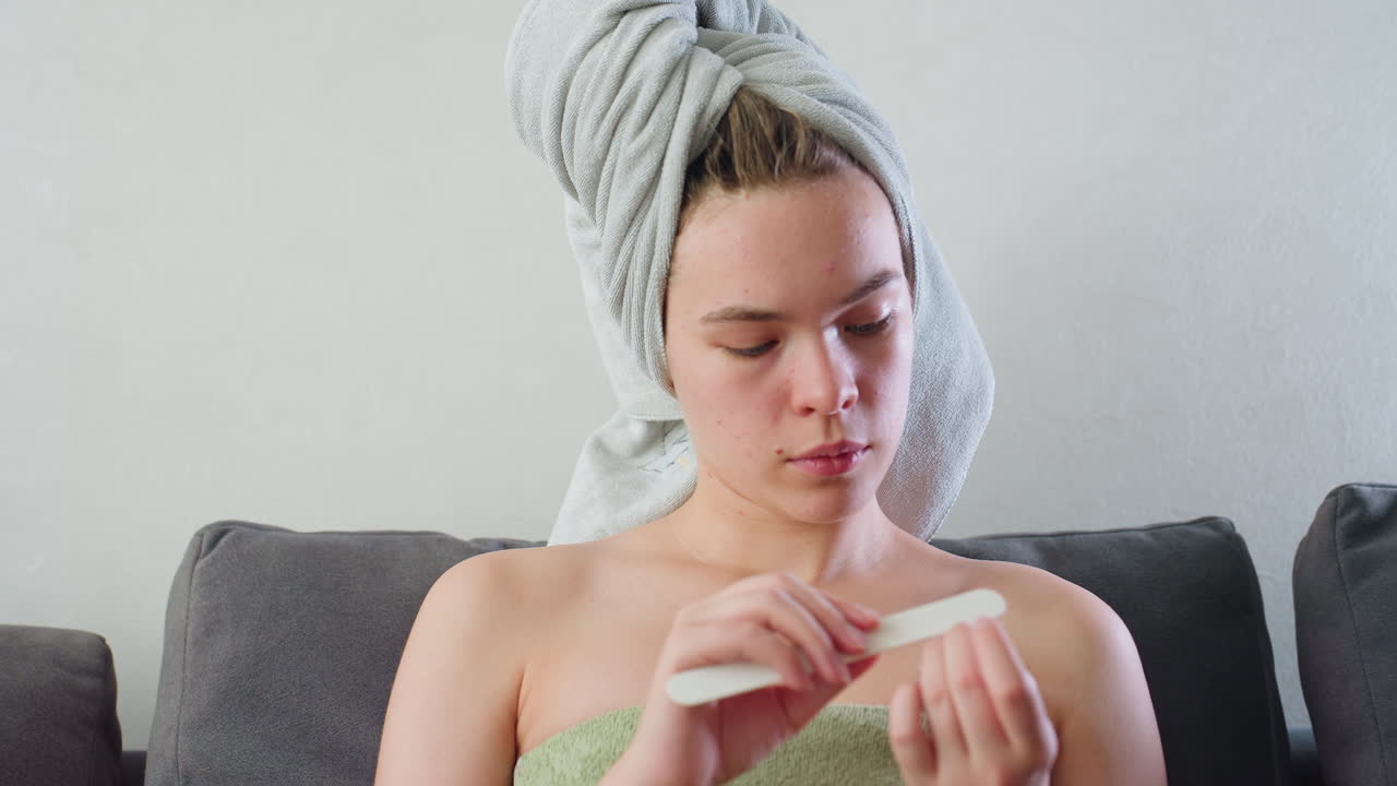 Close view of young woman sitting on couch with towel wrapped around head and green towel over chest, carefully filing nails while relaxed in soft indoor lighting, face shows natural skin