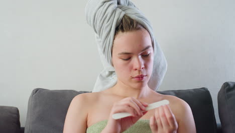 close view of young woman sitting on couch with towel wrapped around head and green towel over chest, carefully filing nails while relaxed in soft indoor lighting, face shows natural skin