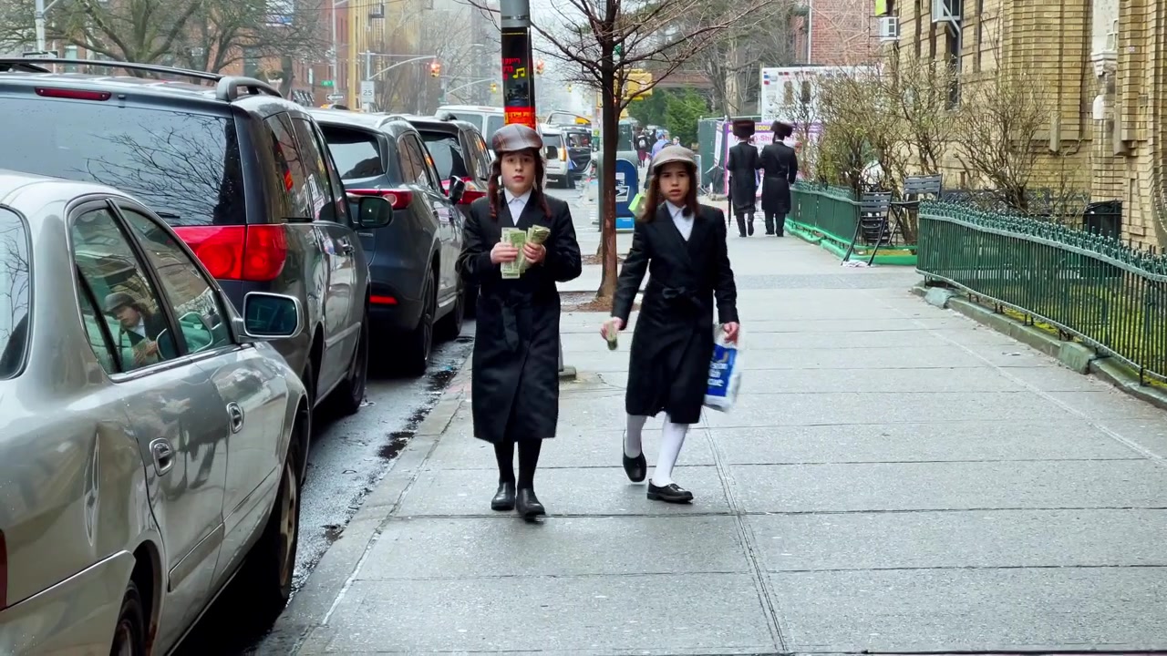 New York, USA, 9 March 2022: Children in black coats walking on Brooklyn sidewalk. Children in black coats walk on wet Brooklyn sidewalk, one holding cash, parked cars behind.