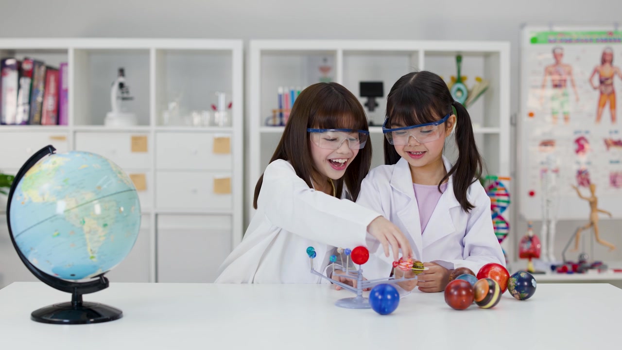 Two young girls in lab coats and safety goggles examine a solar system model.