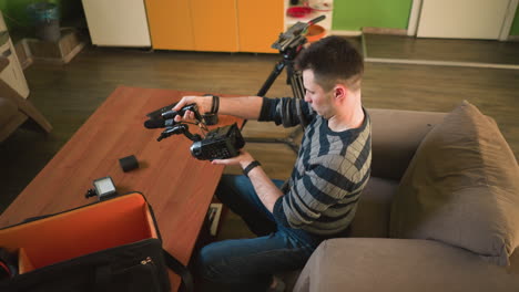 man in striped sweater reaching for camera gear from bag. on wooden table: camera lenses, microphone, smartphone, and other equipment. bright studio space with green walls and cozy atmosphere
