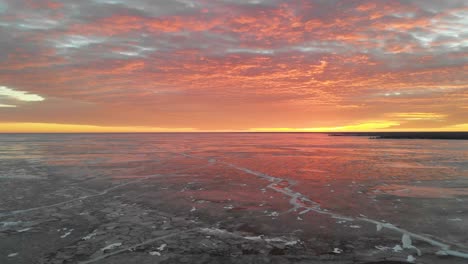 drone rising vertically above a large frozen lake looking at a bright red sunrise that colors the sky and is reflected on the ice.