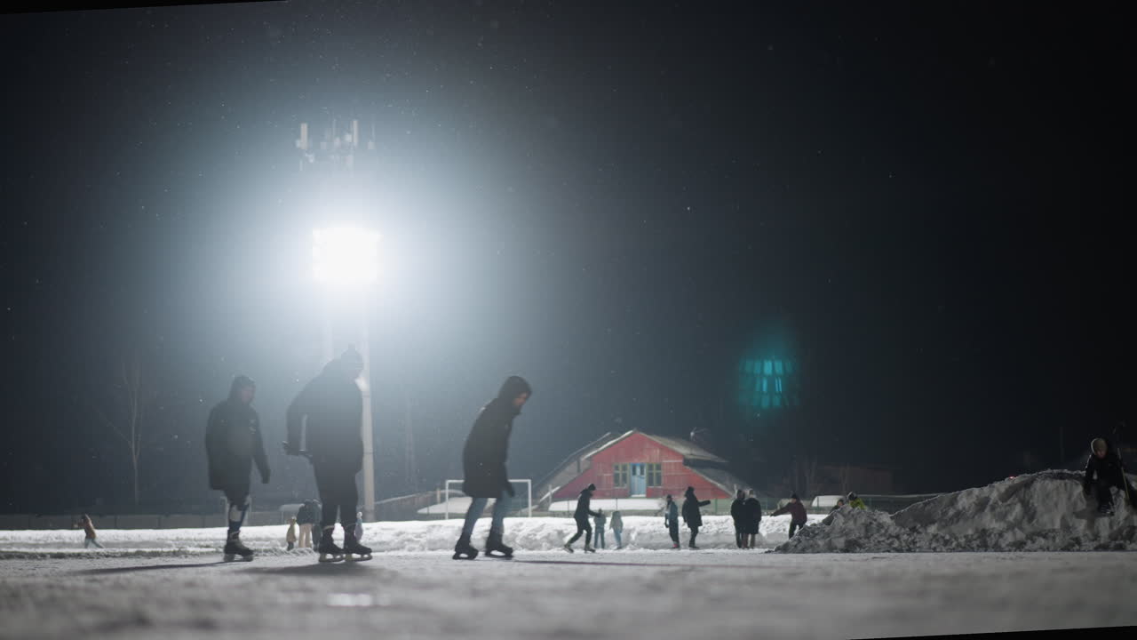 Group of skaters gliding across snow covered ice rink under glowing stadium light at night, surrounded by falling snowflakes and distant figures engaging in fun winter activity