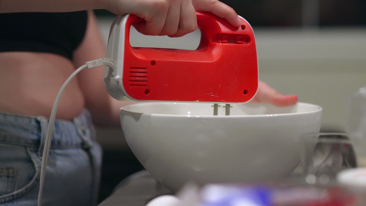 부엌에 있는 하얀 그릇에 치즈케이크 재료를 섞기 위해 빨간 손 믹서를 사용하는 여자