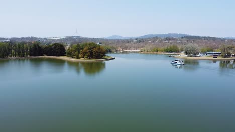 Fliegen-In-Richtung-Des-Parlamentsgebäudes-Von-Canberra,-Während-Ein-Touristenboot-Auf-Dem-See-Kreuzt