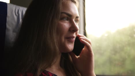 Brown-haired-young-girl-travels-by-modern-train.-Next-to-the-window.-Speaks-on-the-mobile-phone.-Smiling.-Daytime