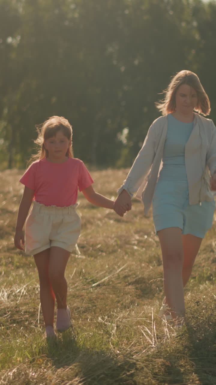 Two sisters walk with child in farmland on sunny day, hair and clothing fluttering in wind, child holding her aunt's hand as they stroll, enjoying fresh air and nature with green trees in background