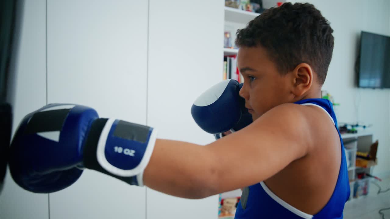 Determined young boy wearing blue boxing gloves, practicing powerful punches on black punching bag while developing athletic skills in home gym environment