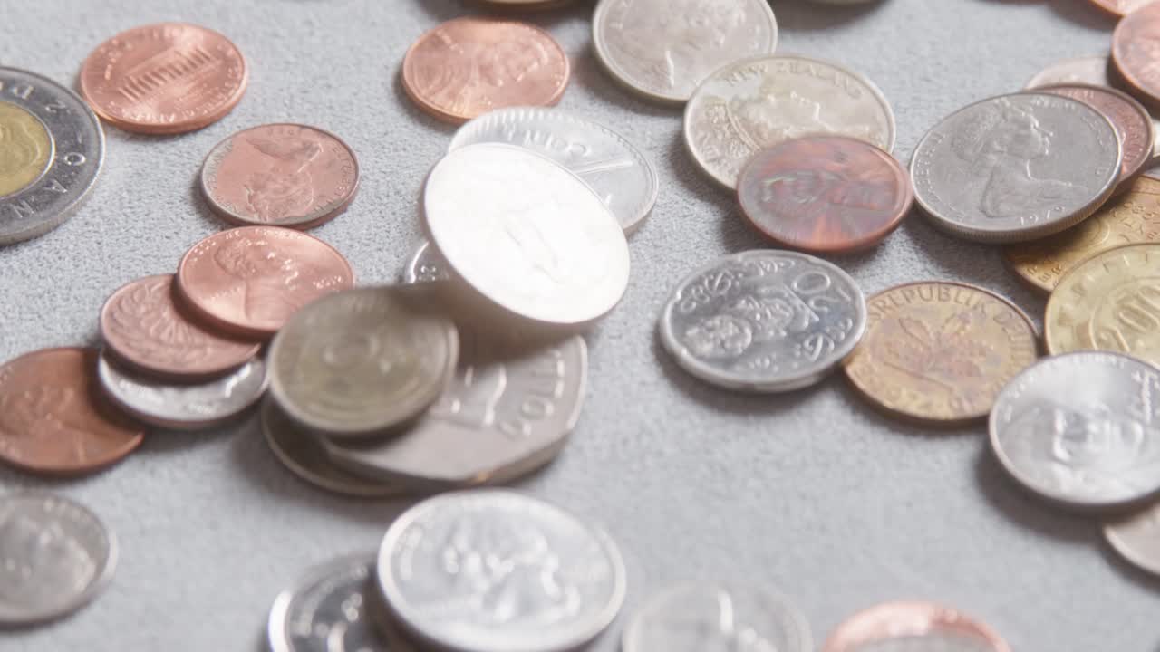 Assorted international coins falling onto a material cloth surface from various countries and denominations, different sizes, values, colours. Slow camera movement past the coins, extreme close up