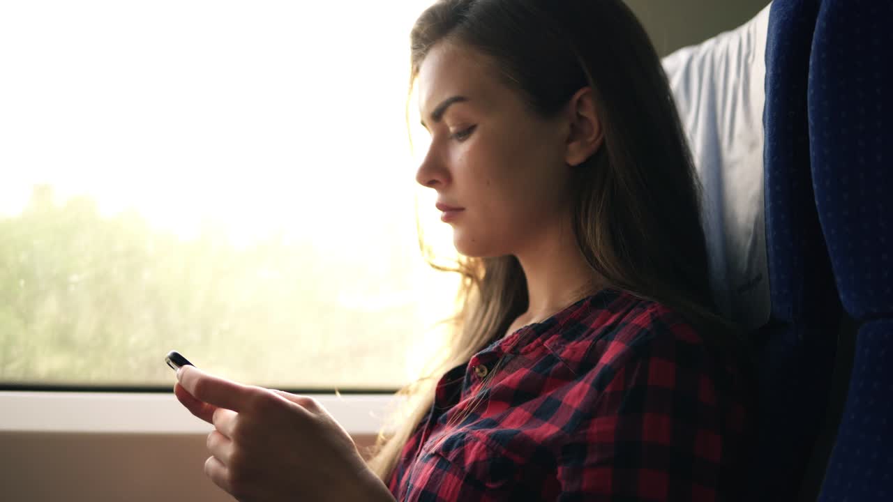mujer atractiva con camisa a cuadros va en tren, sentada junto a la ventana y mirando seriamente su teléfono móvil
