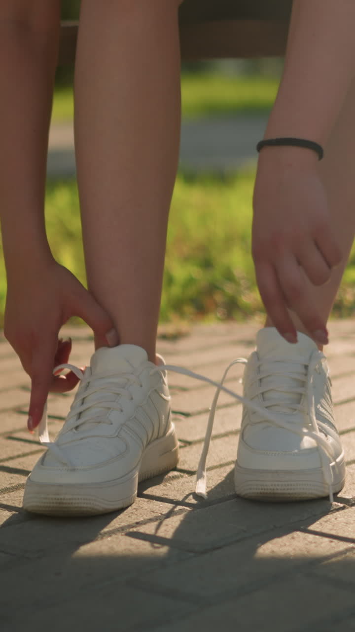 왼쪽 스니커의 신발을 풀고 밖의 벤치에 앉아있는 여자의 다리 시각, 배경의 초록색, 밝은 빛은 보행길에 부드러운 그림자를 만니다.