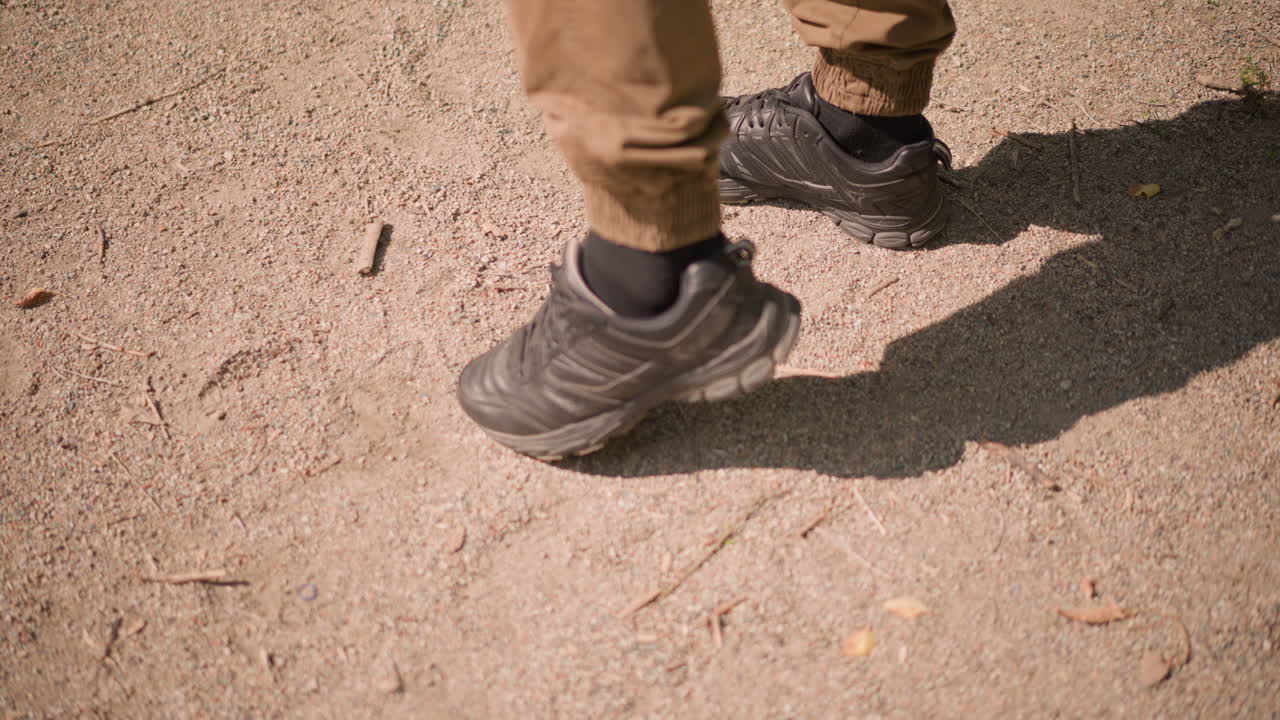 Man Walking Across Dusty Path Leaving Bottle Behind, Low Angle Of Shoes And Shadow Emphasize Steady Stride, Khaki Trousers And Casual Pace Suggest Commuter Or Park Visitor, Natural Light Highlights