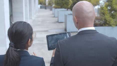 back view of two business colleagues discussing project and watching presentation on tablet while walking at office building