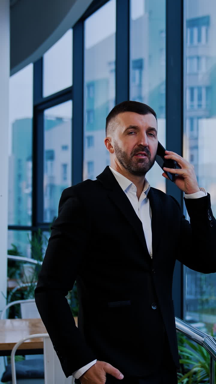Serious man in suit stands holding phone at his ear. Businessman has a phone talk looking at the window. Vertical video