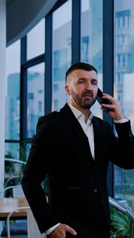 serious man in suit stands holding phone at his ear. businessman has a phone talk looking at the window. vertical video