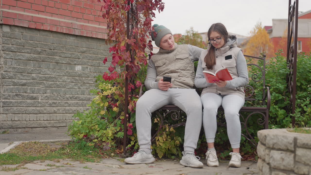 Newly married couple seated outdoors on garden bench in cool breeze as man holds phone and lovingly brings wife close while she reads book, surrounded by greenery, flowers, brick walls
