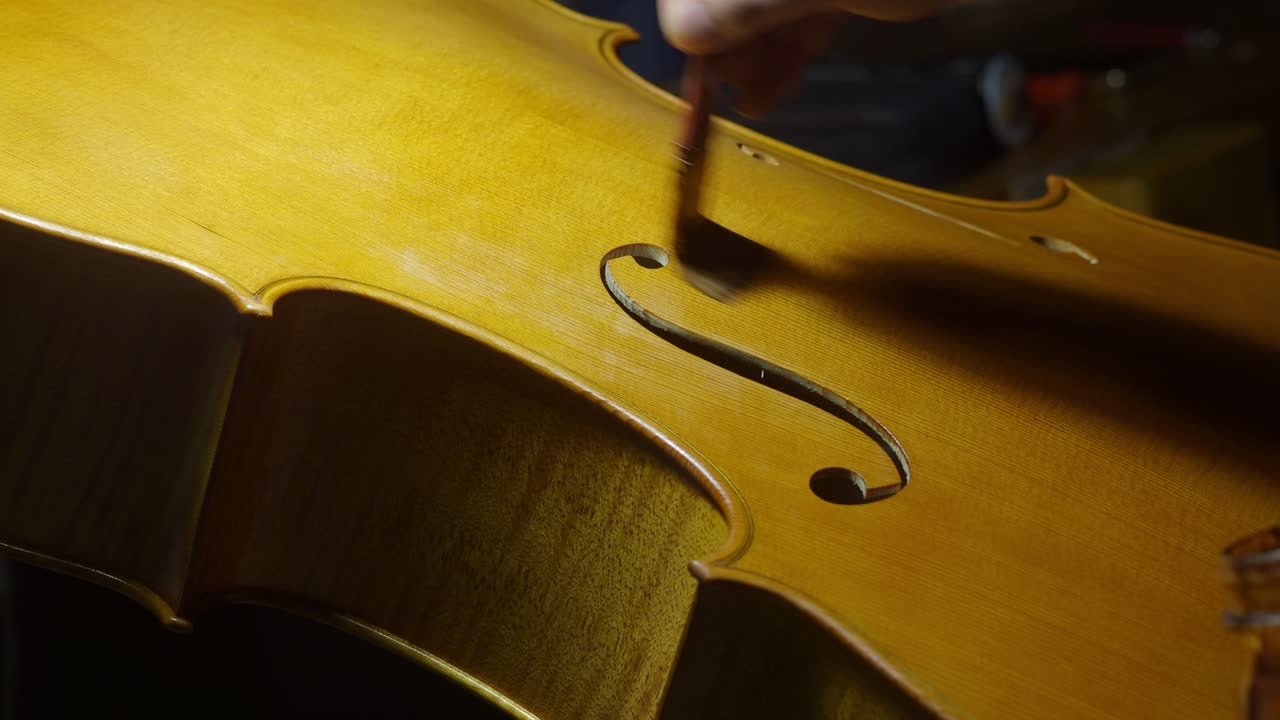 Detailed close-up shows a craftsman carefully applying varnish to the top plate of a cello, highlighting the elegant curves and f-holes in a traditional Cremona luthier’s workshop
