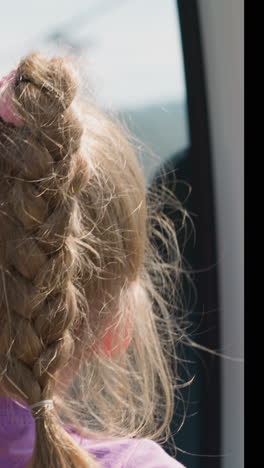 cute little girl with ponytail plait turns head to look at green valley riding cabin of ropeway at highland close backside view slow motion