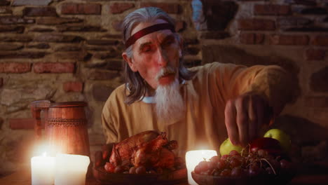 un señor anciano rasga una pierna de pollo en una fiesta medieval. un aristócrata de pelo gris con barba de cabra come carne horneada en la mesa del comedor de la taberna. un rico regalo para el huésped de la posada