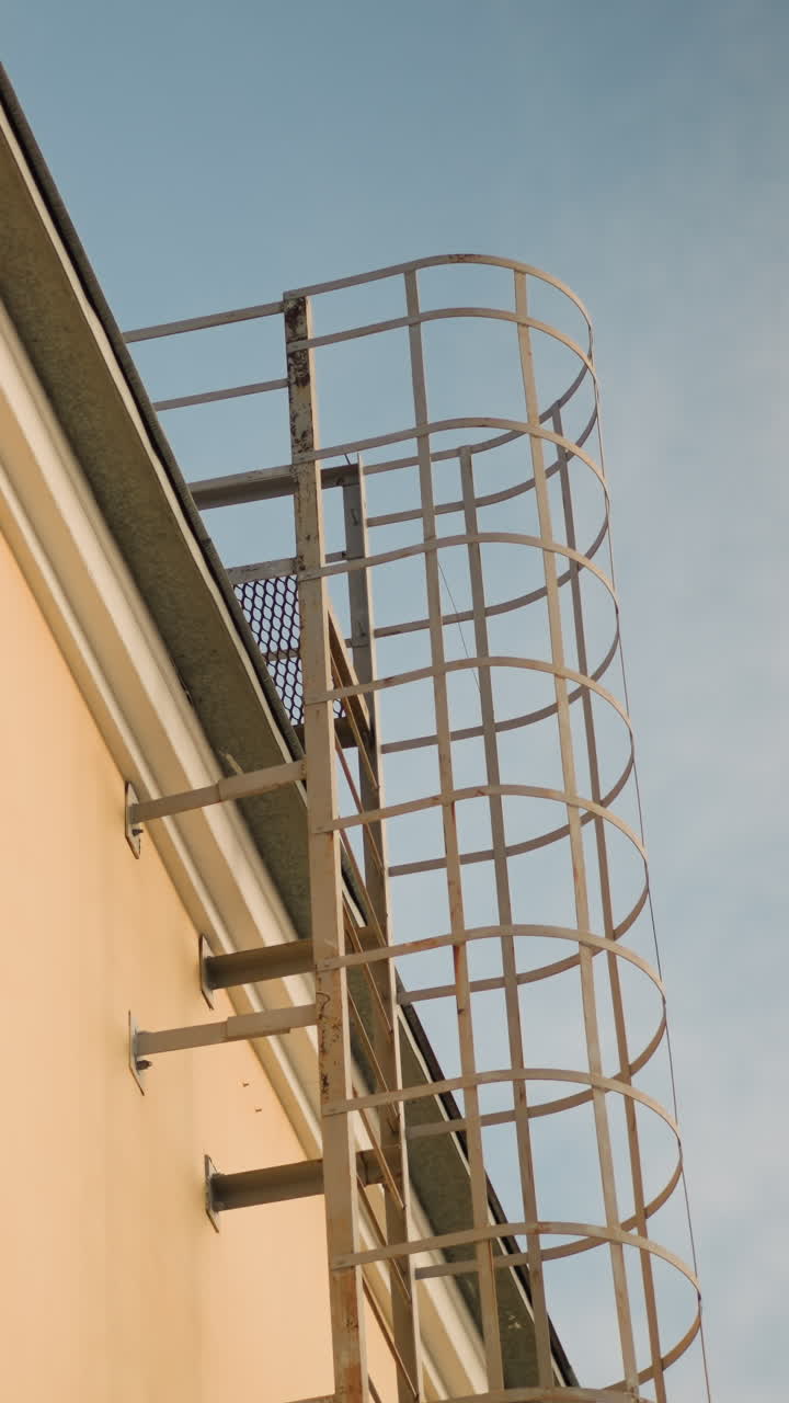 escalera de seguridad exterior con jaula en edificio, jaula metálica unida a una fachada beige bajo un cielo azul despejado; la perspectiva vertical enfatiza la altura y la rejilla protectora, soportes robustos y línea de tejado