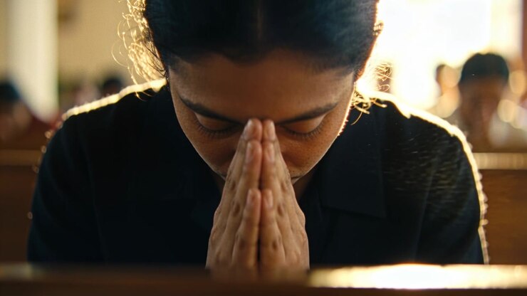 Praying woman in dark collared top lowering head at church, holding clasped hands on wooden pew