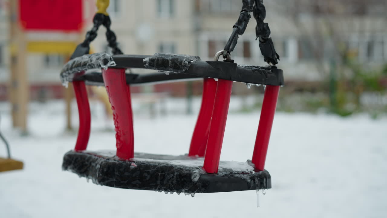 Jäätynyt lasten keinu, jään ja jääpuikkojen peitossa, roikkuu keltaisilla köysillä punamustasta rungostaan ja keinuu hiljaa lumista leikkikenttää ja sumeaa rakennustaustaa vasten
