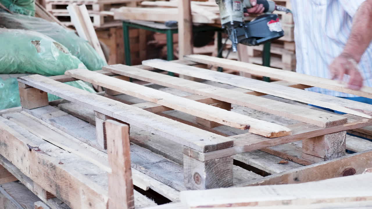 trabajador de un hombre en una fábrica de madera usando una pistola de clavos para hacer un pallet de madera