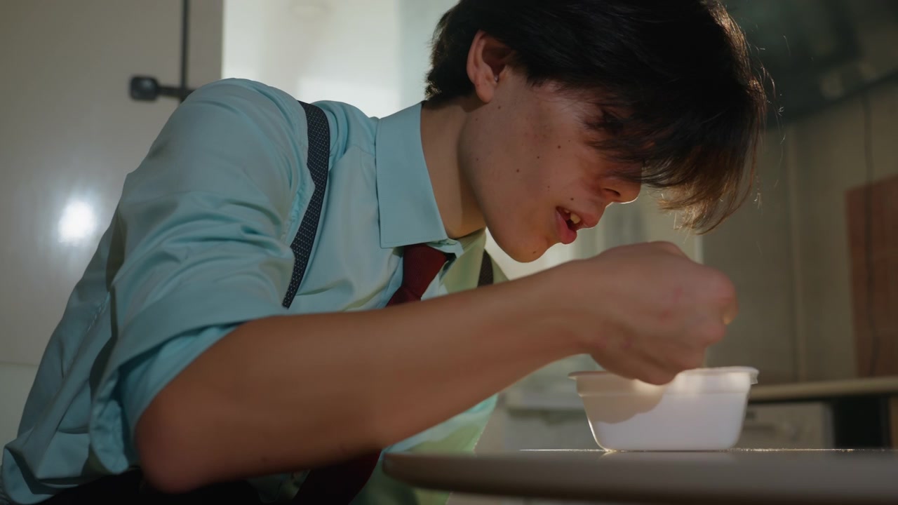 キッチンで湯気の立つスープを食べるアジア人男性。シャツ、ネクタイ、サスペンダーを着用した若いアジア人のインターンが、スプーンの一口に集中している。窓から差し込む朝日に湯気が照らされ、熱い味を捉えるシネマティックなクローズアップ。