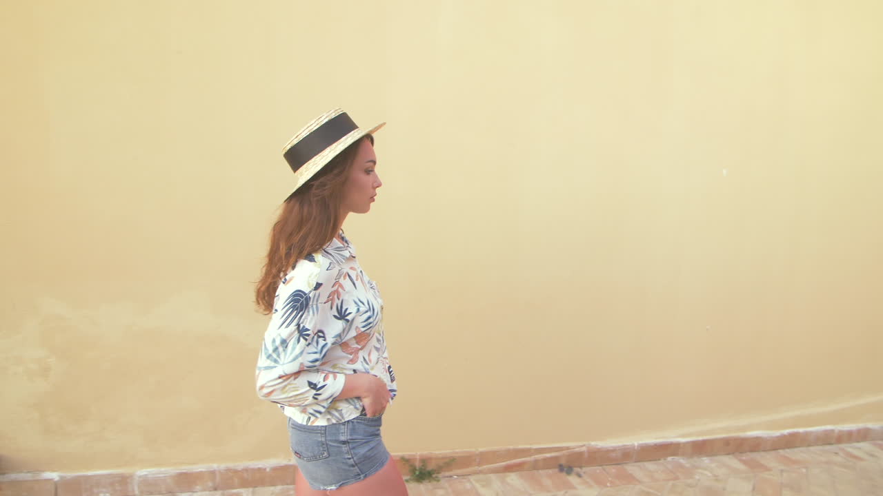 una mujer joven con un sombrero de paja y una camisa floral camina frente a una pared amarilla.