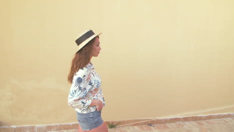 a young woman in a straw hat and floral shirt walks in front of a yellow wall.