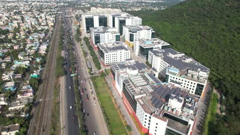 Toma-Aérea-De-Edificios-Corporativos-Cerca-De-Una-Colina-Llena-De-árboles-Y-Una-Carretera-Llena-De-Autos-Que-Pasan