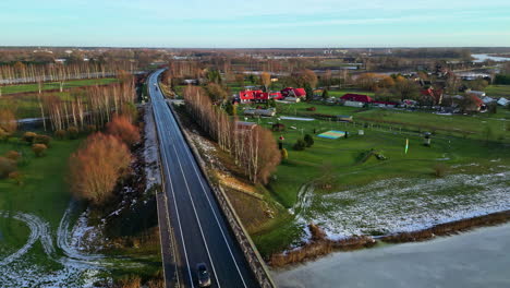 road winding through a rural landscape with sparse snow, vehicles, and surrounding greenery, at dusk