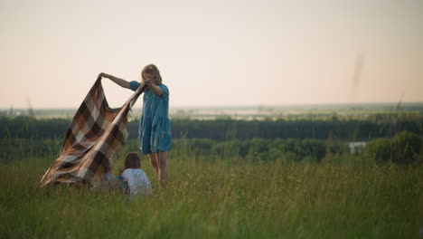 a woman in a blue gown holds a plaid scarf as the wind gently blows it in a grassy field overlooking a scenic river. two happy children in white shirts bend down as she tries to cover them