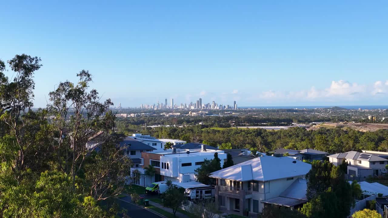 Drone si eleva sopra case residenziali e alberi rivelando lo skyline di una città distante sotto cieli blu.