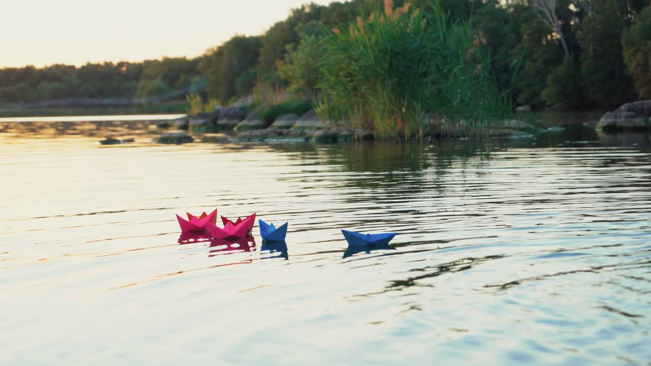 Different colored paper ships swim in a quick river surface on the natural background with green trees. Origami boats in bright colors on a lake at sunset